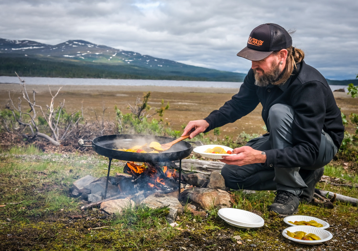 Ola Johansson från Taktältarna lagar mat på stekhäll över öppen eld. Sjö och fjäll i bakgrunden